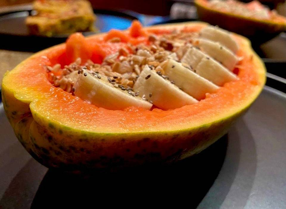 Half of a papaya fruit with orange flesh, filled with sliced pieces of white fruit, sprinkled with black seeds, on a black plate.