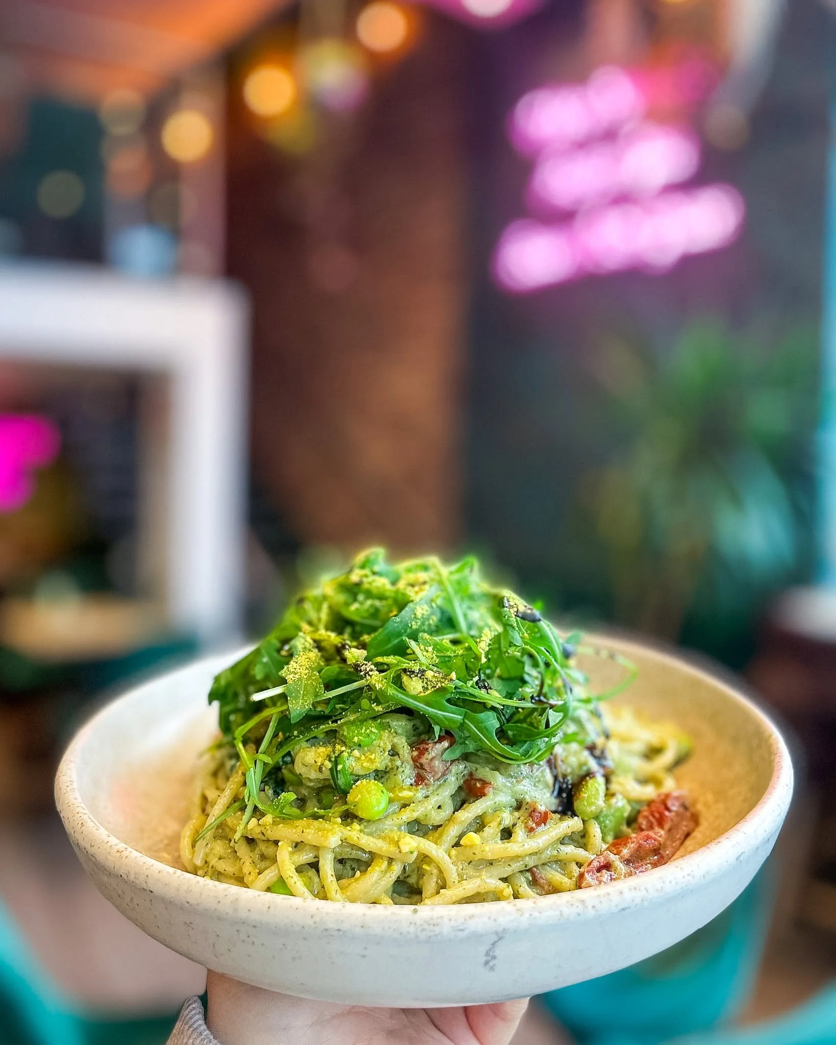 A bowl of spaghetti pasta topped with fresh arugula, peas, and tomato sauce, held in a person's hand.