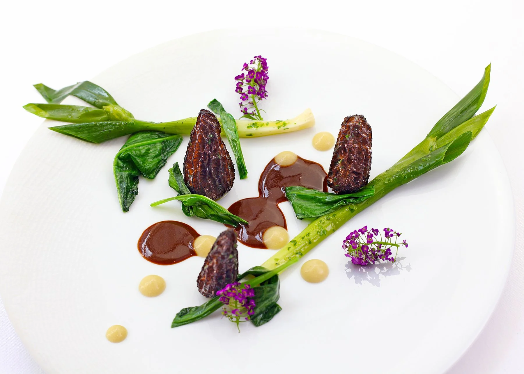 Elegant plated gourmet dish with green vegetables, purple edible flowers, dark sauce, and small white dollops on a white round plate.