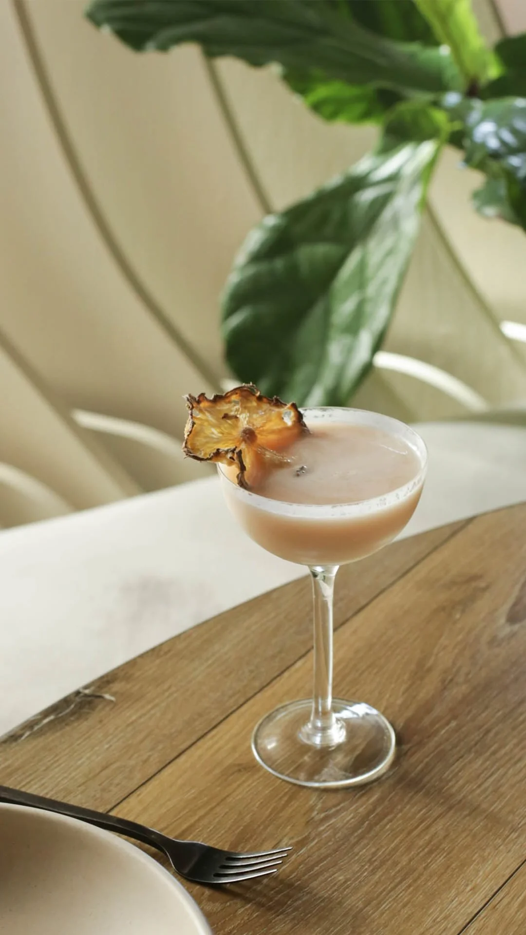 A cocktail in a coupe glass garnished with a dried pineapple slice, placed on a wooden table with a fork and a bowl in the foreground, and green leaves in the background.