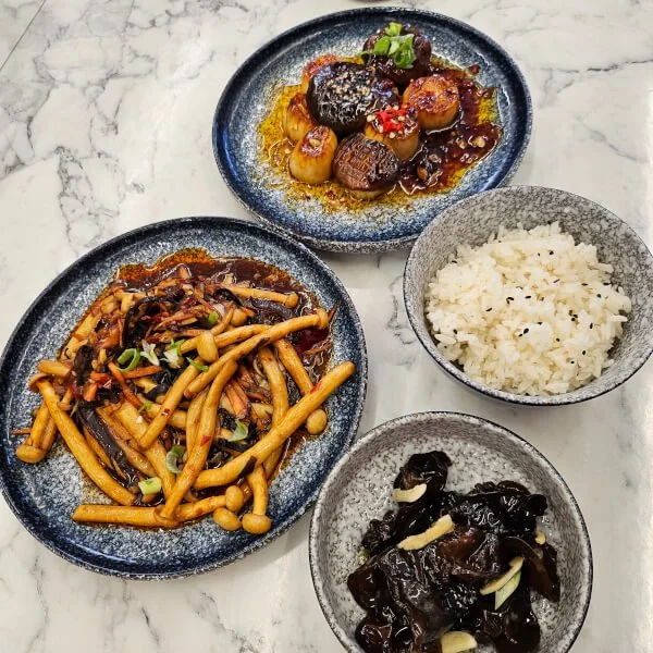 Assorted Chinese dishes on a marble table: a bowl of white rice, a plate of stir-fried mushrooms and vegetables, and a plate of braised meat with vegetables garnished with chopped green onions.