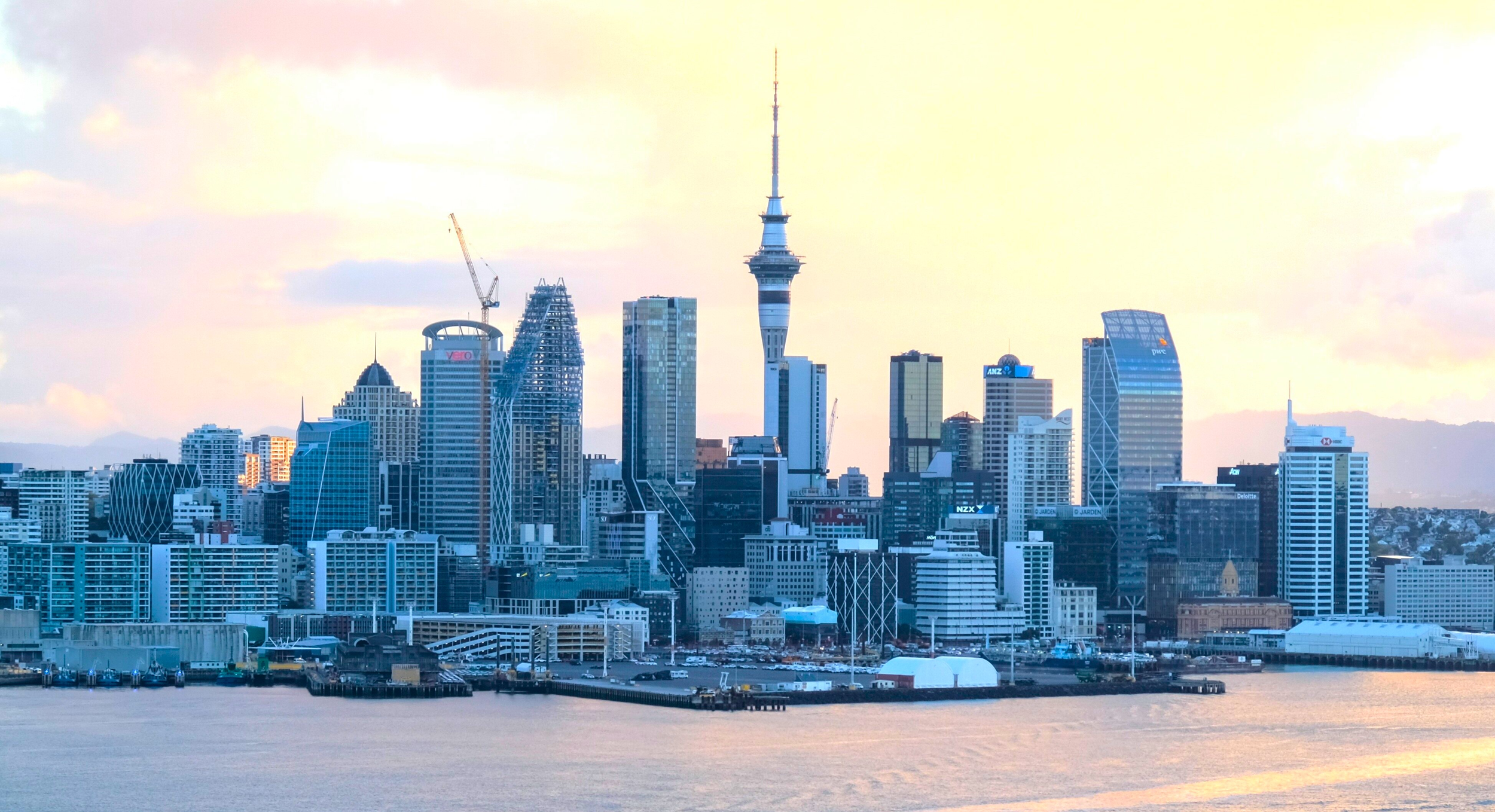 City skyline with modern skyscrapers and a prominent tower, near water with a waterfront in the foreground. Cloudy sky with warm sunlight.