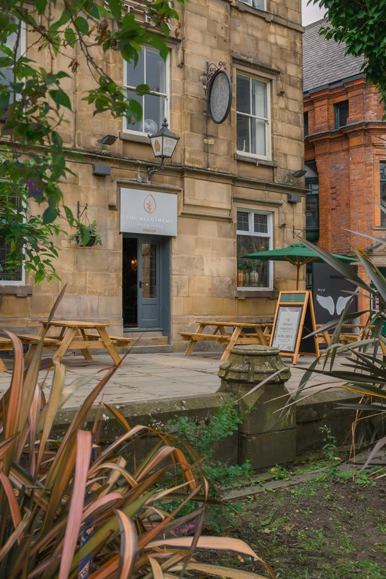 Exterior view of a café or restaurant named The Allotment with outdoor seating, a green umbrella, and a chalkboard sign, in a historic brick building with trees and plants in the surrounding area.