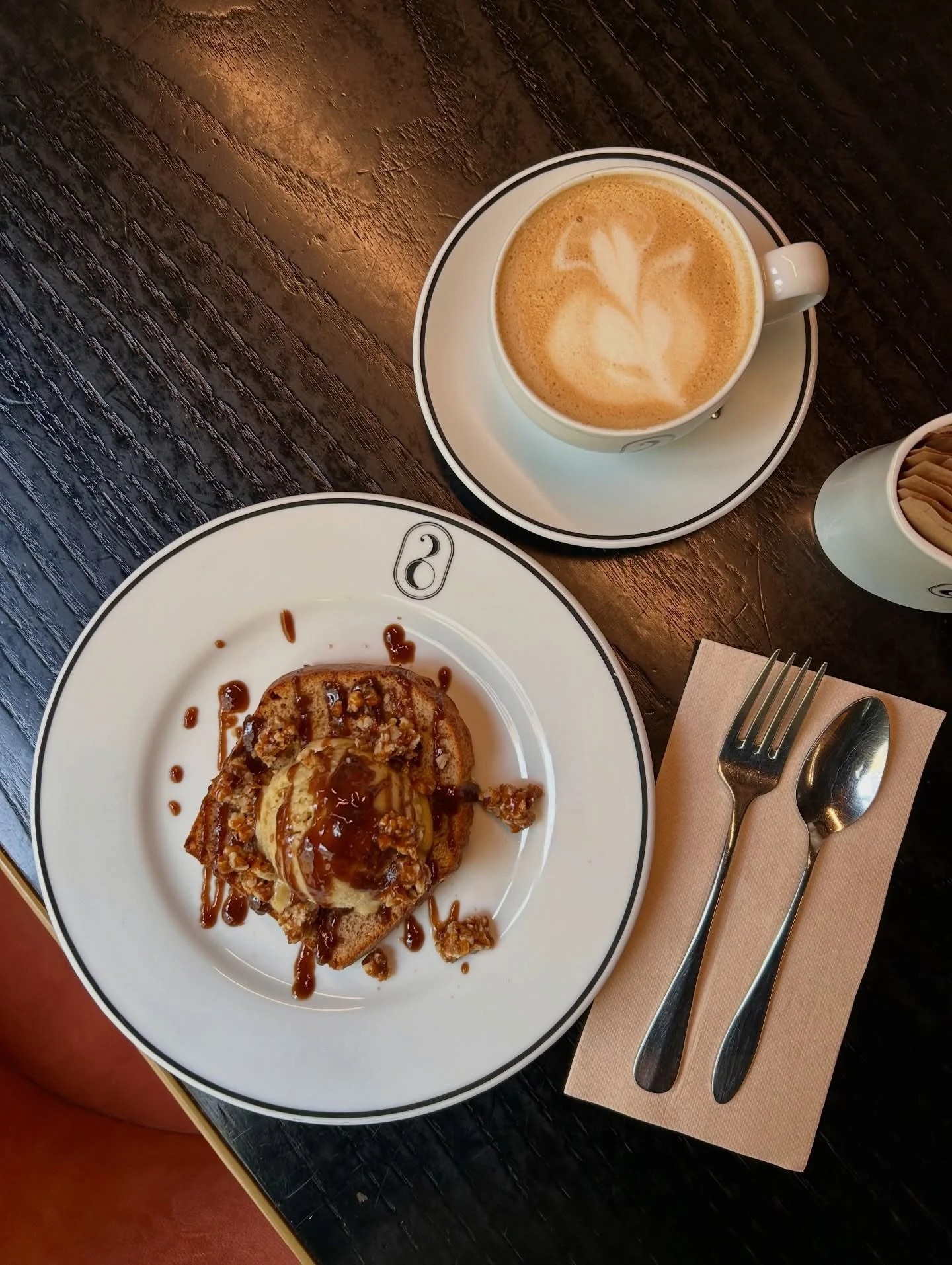 A plate with a cinnamon roll drizzled with caramel sauce, a cup of latte with heart-shaped foam art, and a container with packets of sugar or creamer on a dark wooden table, with a fork and spoon on a napkin.