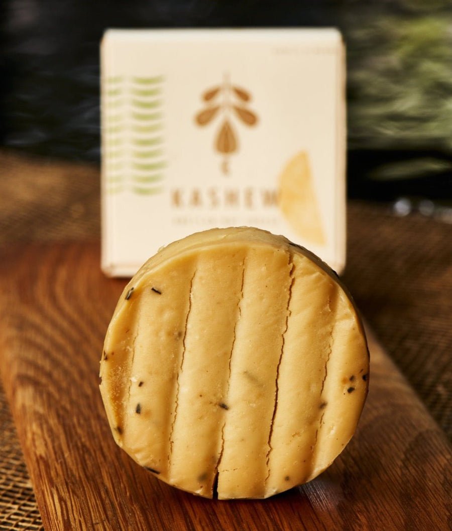 A round wheel of cheese with visible herbs, placed on a wooden surface, with a box featuring a bee and herbs in the background.
