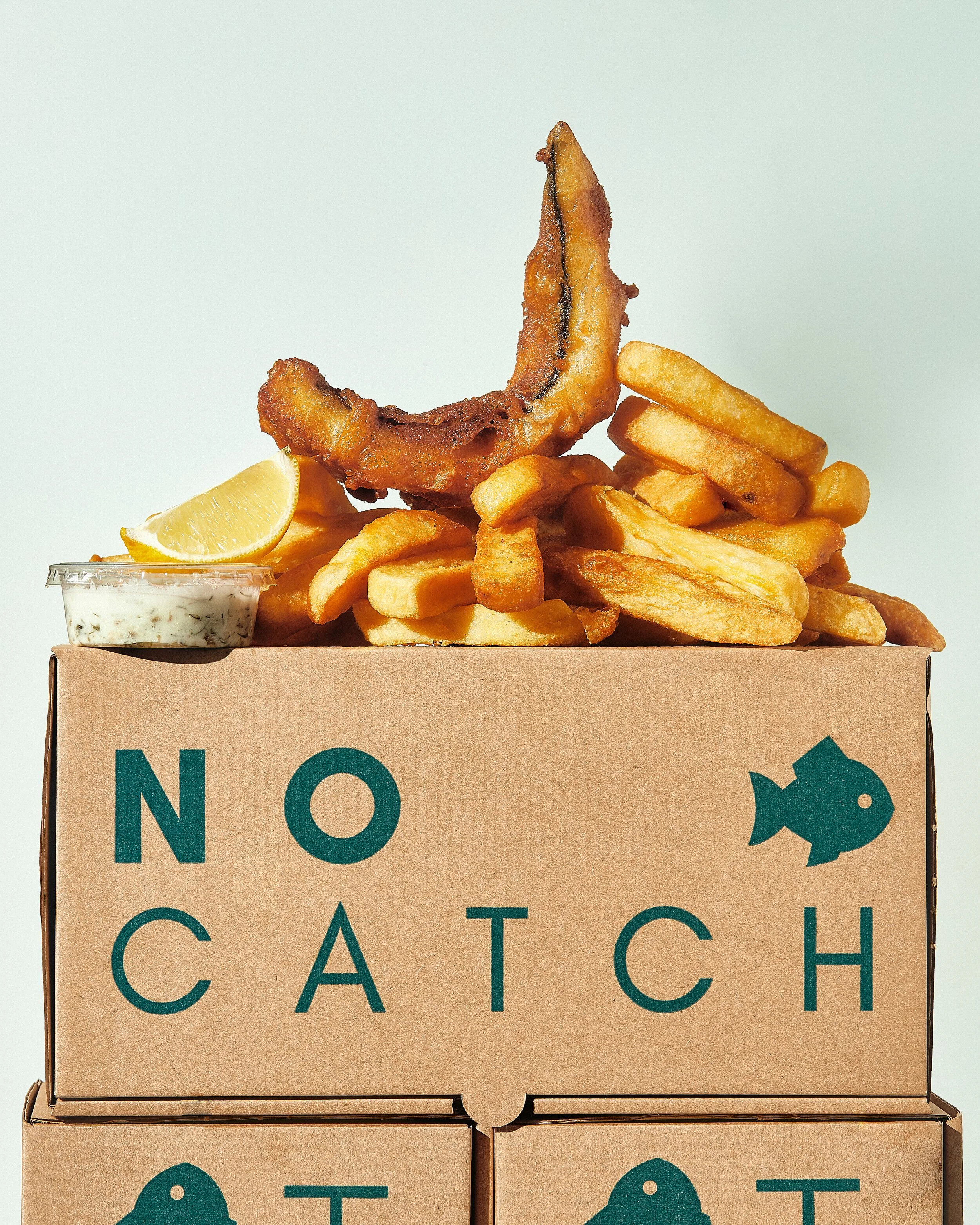A cardboard box with the words 'NOT CATCH' printed on it, containing a large serving of French fries, a fried fish fillet, a lemon wedge, and a small container of tartar sauce.