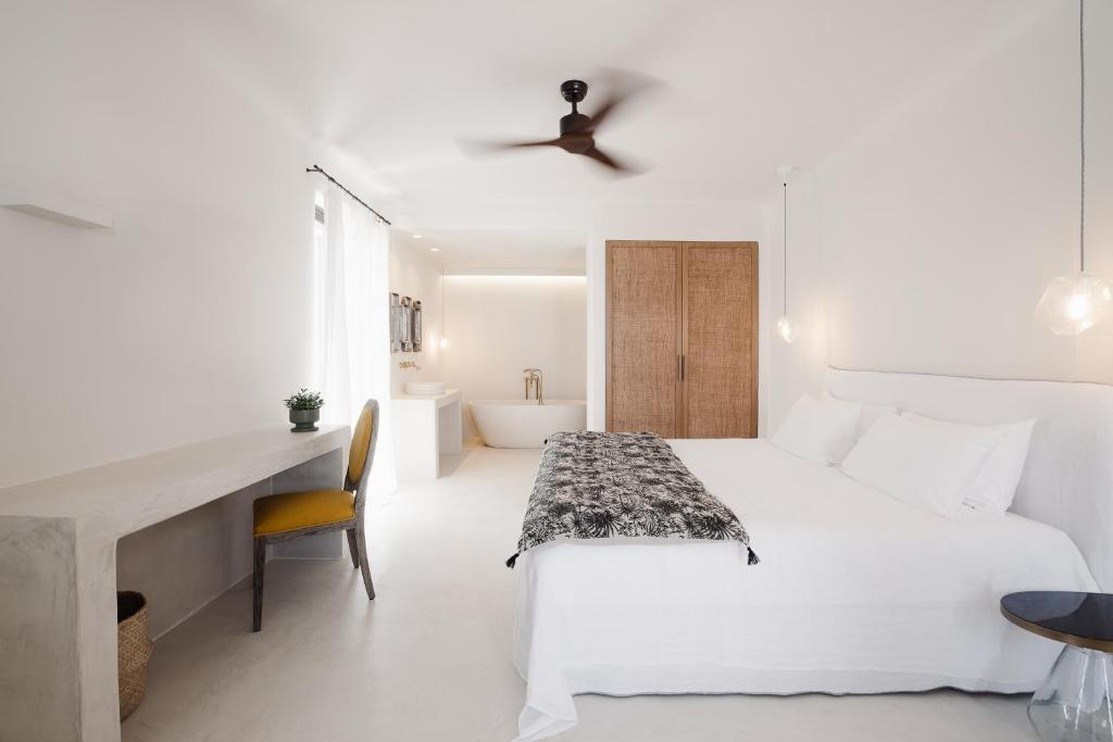 Bright, minimalist bedroom with white walls, a large bed with white linens, bedside pendant lights, a wooden wardrobe, a yellow chair at a marble desk, a small plant, and a bathtub in the background by a window with sheer curtains.