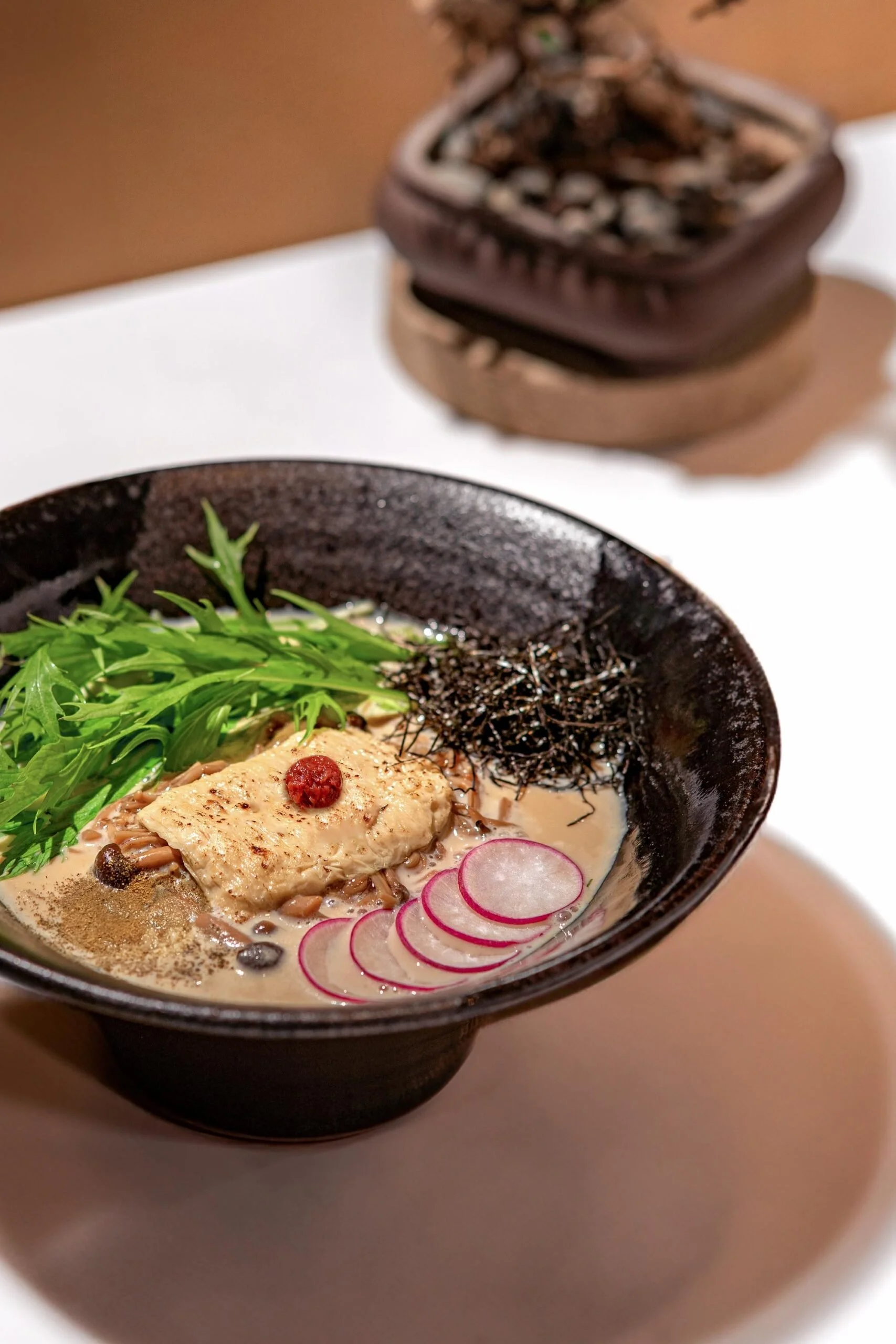 A bowl of ramen with sliced radish, fresh greens, and a piece of chicken topped with a red chili pepper.