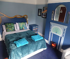 A cozy bedroom with a bed covered in a dark teal quilt and green patterned pillows, two folded towels on the bed, a blue nightstand with a book and a small lamp, framed pictures on the wall, a mirror above a white dresser with intricate carvings, and a small red candle holder on the floor.