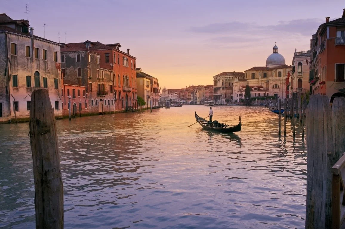 Sunset over a canal in Venice, Italy, with colorful historic buildings on both sides. A gondola with a gondolier in white shirt and dark pants is on the water.