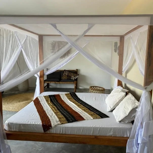 A bedroom with a four-poster canopy bed featuring white mosquito netting, a white mattress with white pillows, and a colorful striped blanket. There is a wooden bench with decorative cushions and a wicker basket in the background.