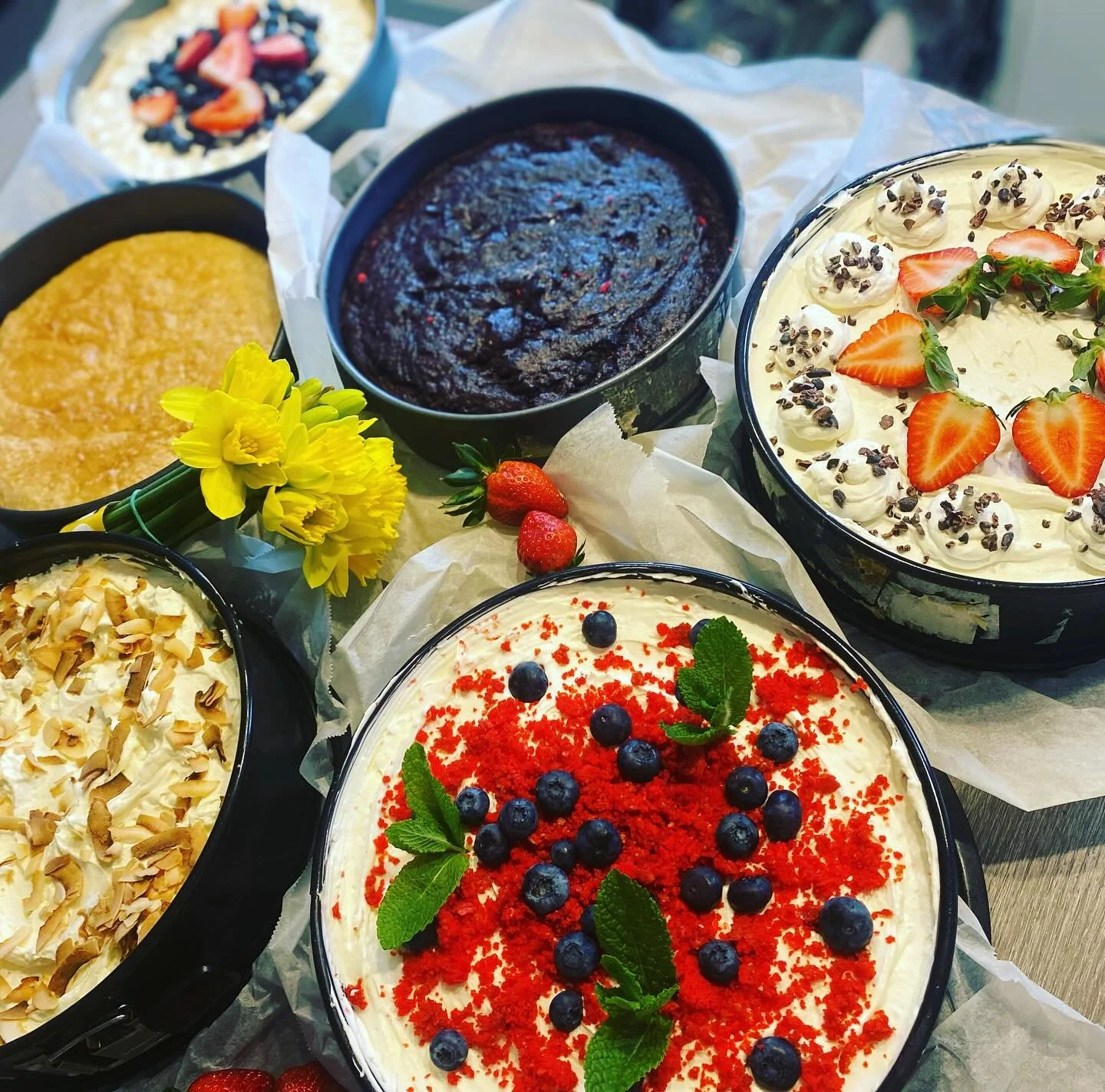 Assorted cakes topped with berries, chocolate shavings, and whipped cream, with fresh strawberries, blueberries, and yellow flowers nearby.