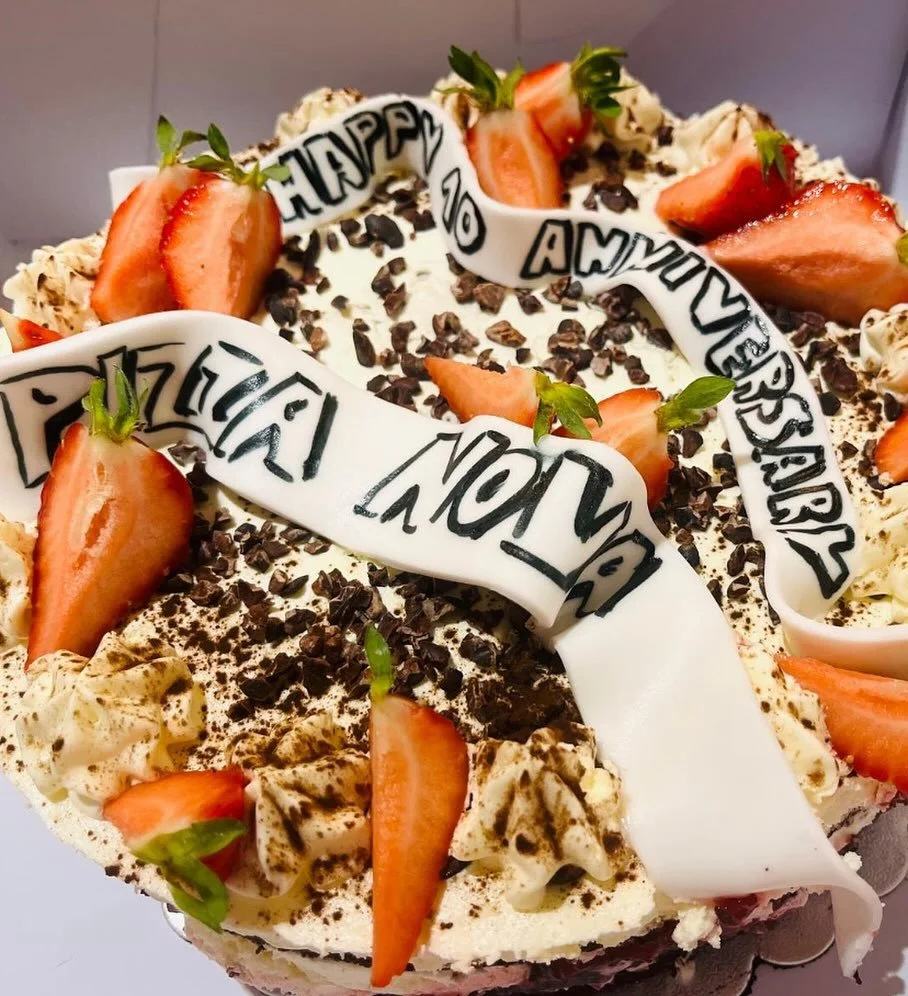 Celebration cake decorated with sliced strawberries, chocolate shavings, whipped cream, and white fondant ribbons with 'Happy Anniversary' written on them.