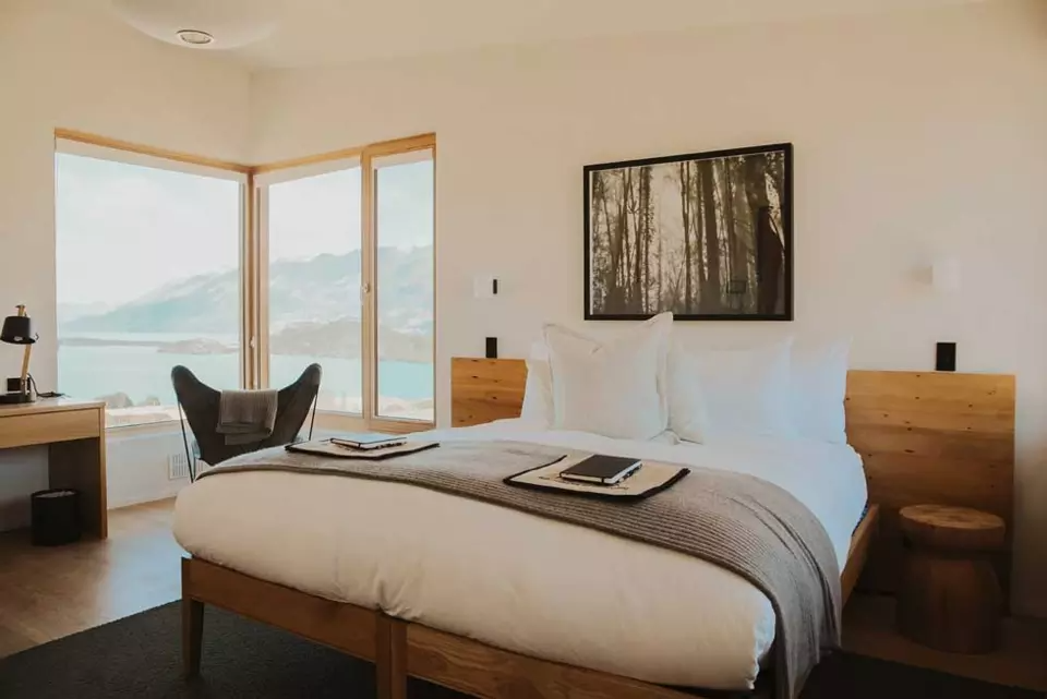 A cozy hotel room with a large bed, pillows, and a gray blanket, overlooking mountains and water through large windows, with a desk, chair, and framed forest picture on the wall.