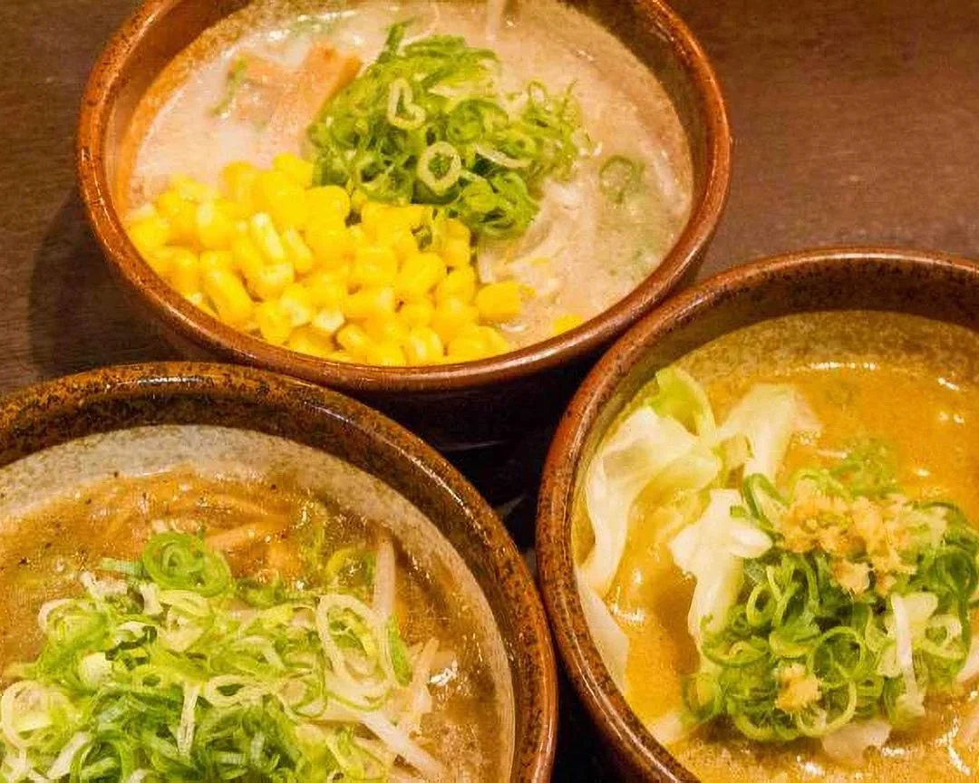 Three bowls of soup with chopped green onions, one bowl with corn, and a bowl of broth with cabbage and green onions.