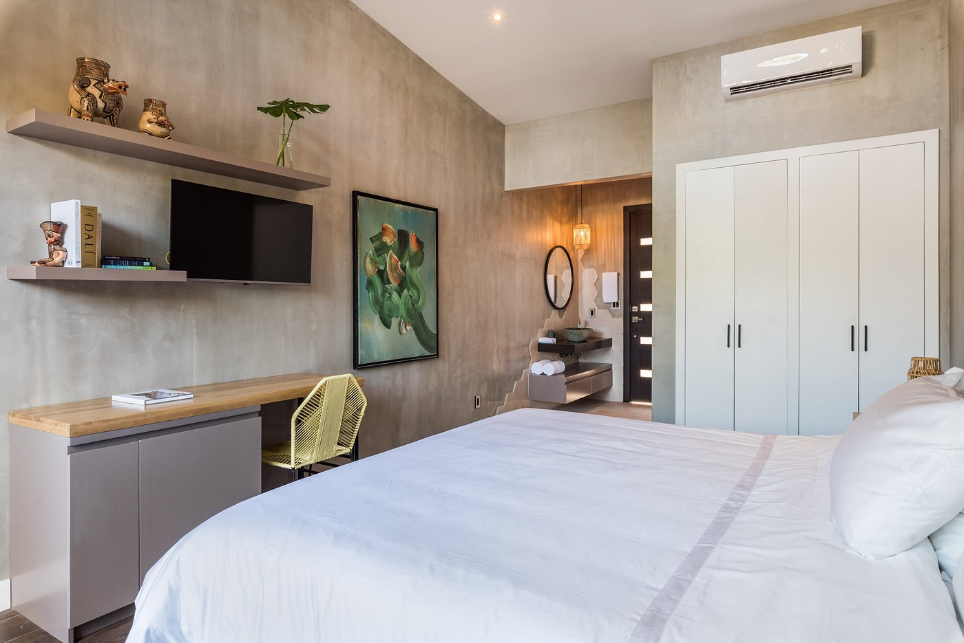 Modern bedroom with a large bed, white bedding, light-colored walls, a flat-screen TV, a desk with a yellow chair, wall-mounted shelves with decorative items, a colorful abstract painting, a mirror, a small bathroom area with a sink and mirror, and a closet with white doors.