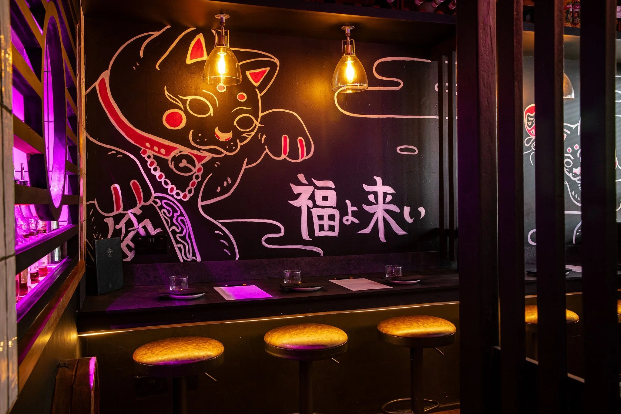 Wall with neon art of a cartoon cat wearing a collar and a pearl necklace, with Japanese characters below. The wall has black background and is illuminated by hanging warm yellow lights. There are bar stools with round seats in the foreground.