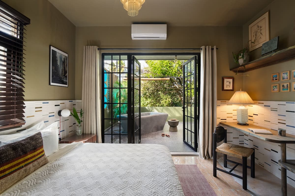 Bedroom with breakfast bar, open glass doors leading to outdoor patio with greenery, wall art, and a window with blinds.