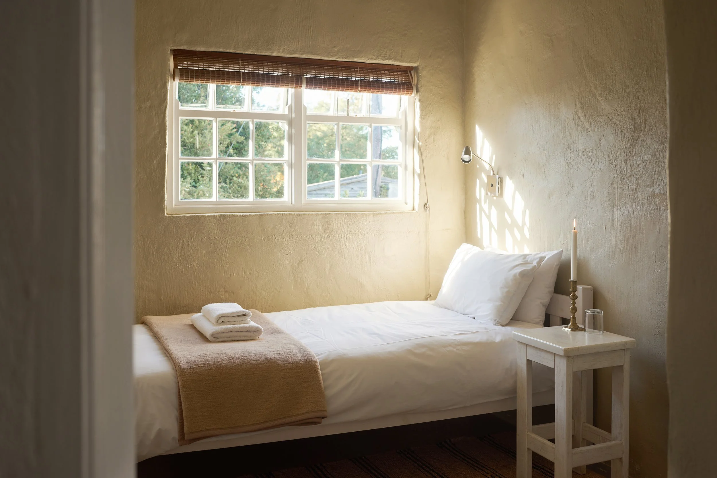 A cozy bedroom featuring a window with a bamboo blind, a white bed with folded towels, a bedside table with a lit candle, a glass of water, a small reading lamp, and natural light streaming in.
