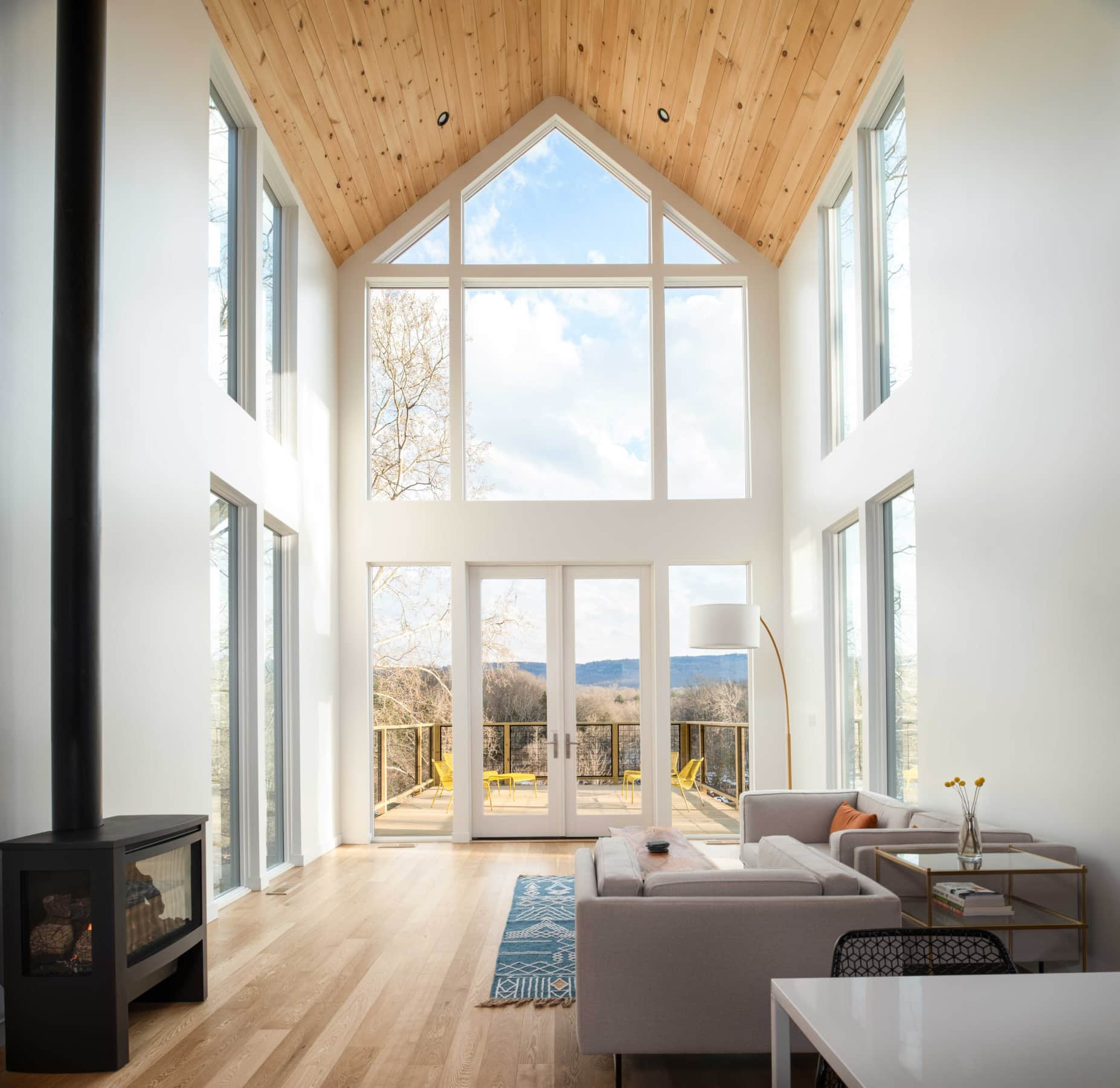 Modern living room with large floor-to-ceiling windows and a high, wooden vaulted ceiling, overlooking a landscape with trees and mountains.