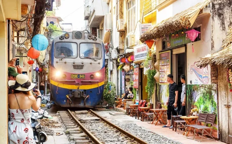 A train passing through a narrow street with colorful shops and cafes. People are taking photos and walking by, with decorative lanterns hanging overhead.