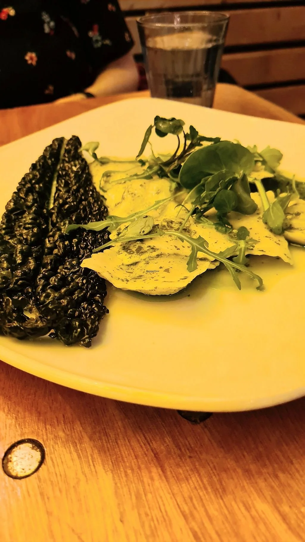 Black rice cake, greens, and sliced yellow zucchini with sauce on a white plate, in a restaurant setting with a wooden table and a glass of water in the background.