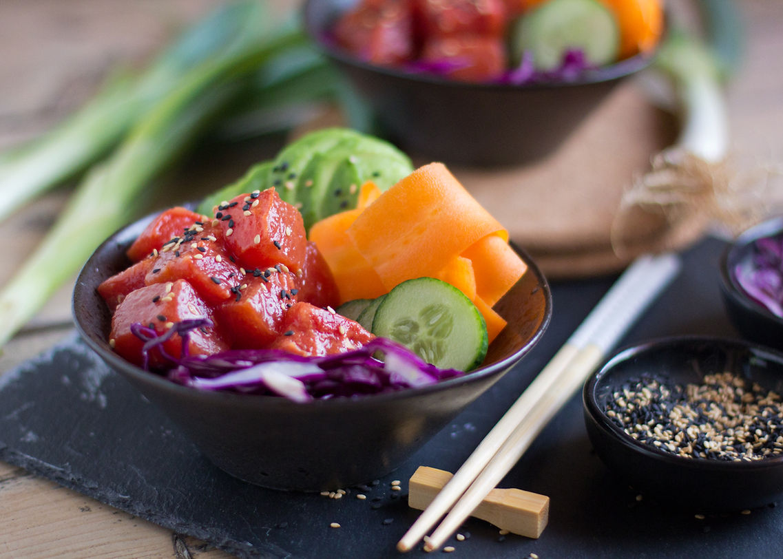 A bowl of poke with raw salmon, cucumber, avocado, shredded carrot, and purple cabbage, topped with black sesame seeds. There are chopsticks beside the bowl and additional bowls of ingredients or sauces nearby on a wooden surface.