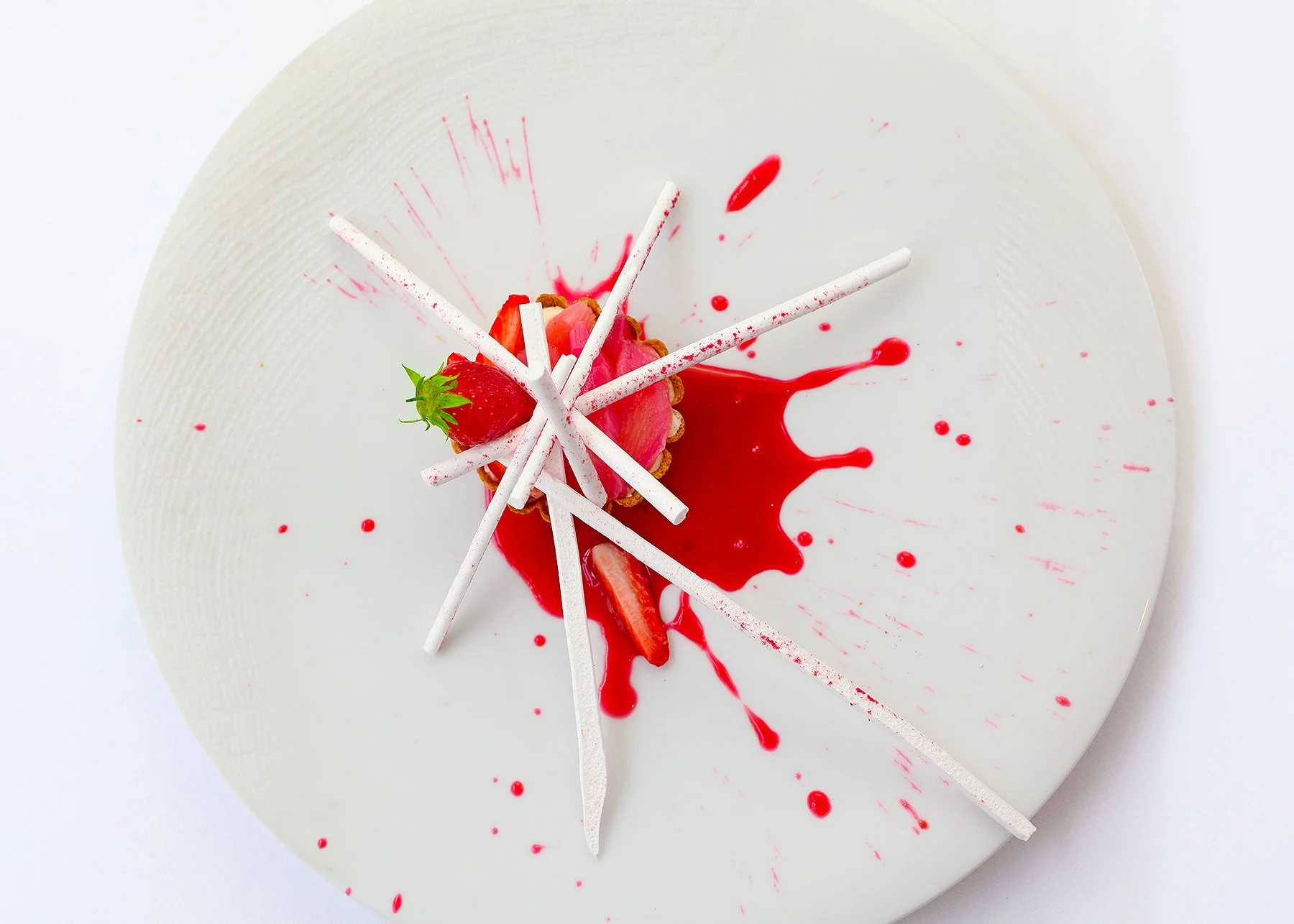 A white, round dessert plate with a strawberry tart decorated with white chocolate sticks and a fresh strawberry. Red sauce and splatters are artfully placed on the plate for presentation.