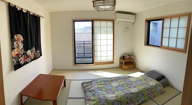 A minimalist bedroom with a futon on a tatami mat, a wooden side table, and shoji-style windows, illuminated by natural light.