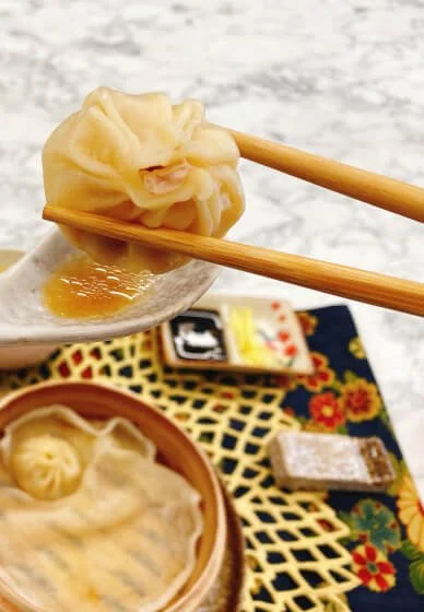 Chopsticks holding a steamed dumpling over a bowl of soup with more dumplings and a tray of condiments visible.
