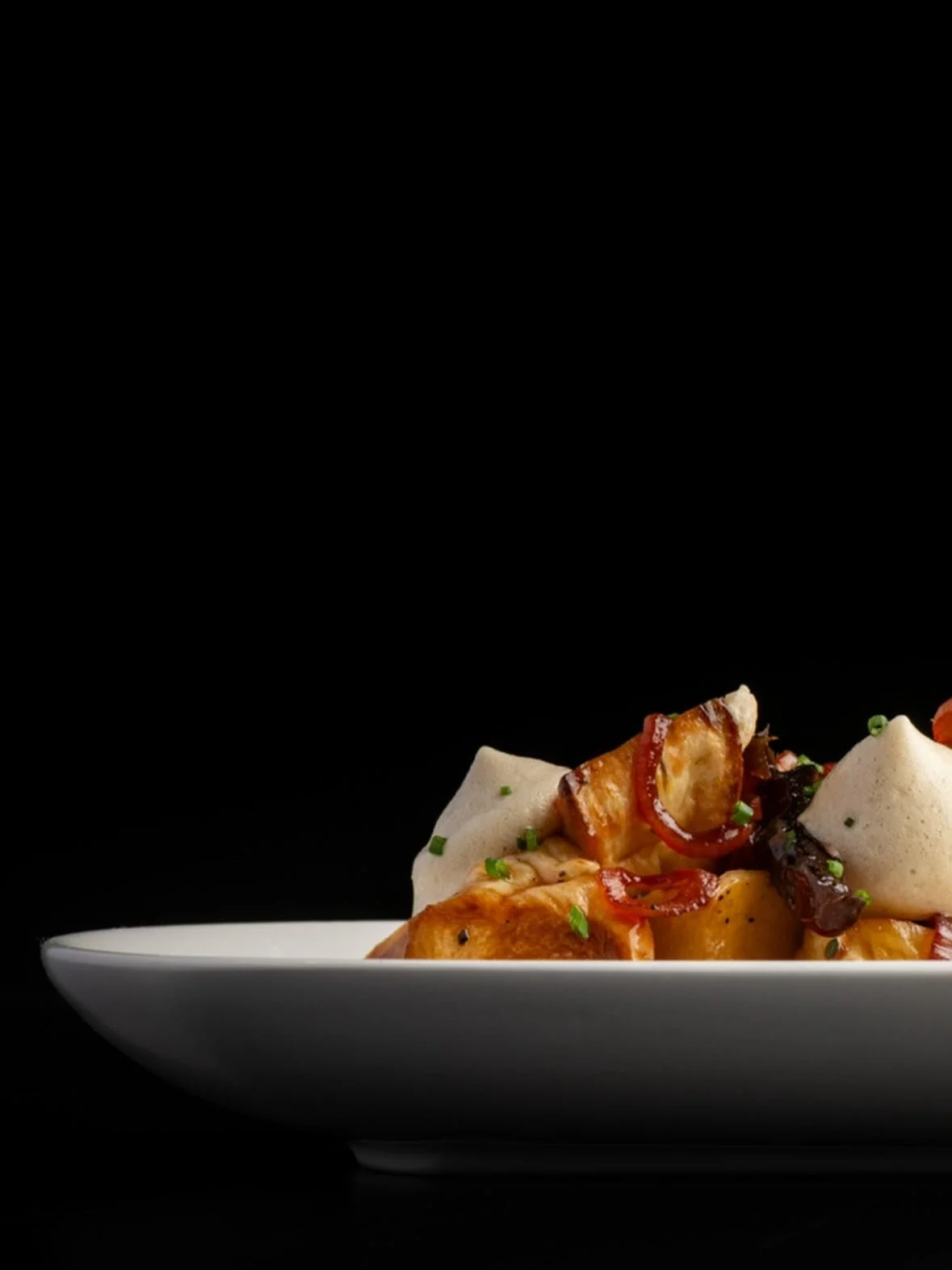 A close-up of a plated gourmet dish with roasted potatoes, thinly sliced red peppers, a dollop of foam, and garnished with chopped green herbs, on a white plate against a black background.