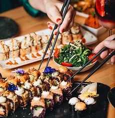 A person using tongs to pick up sushi rolls on a platter, with broccoli and other dishes nearby.