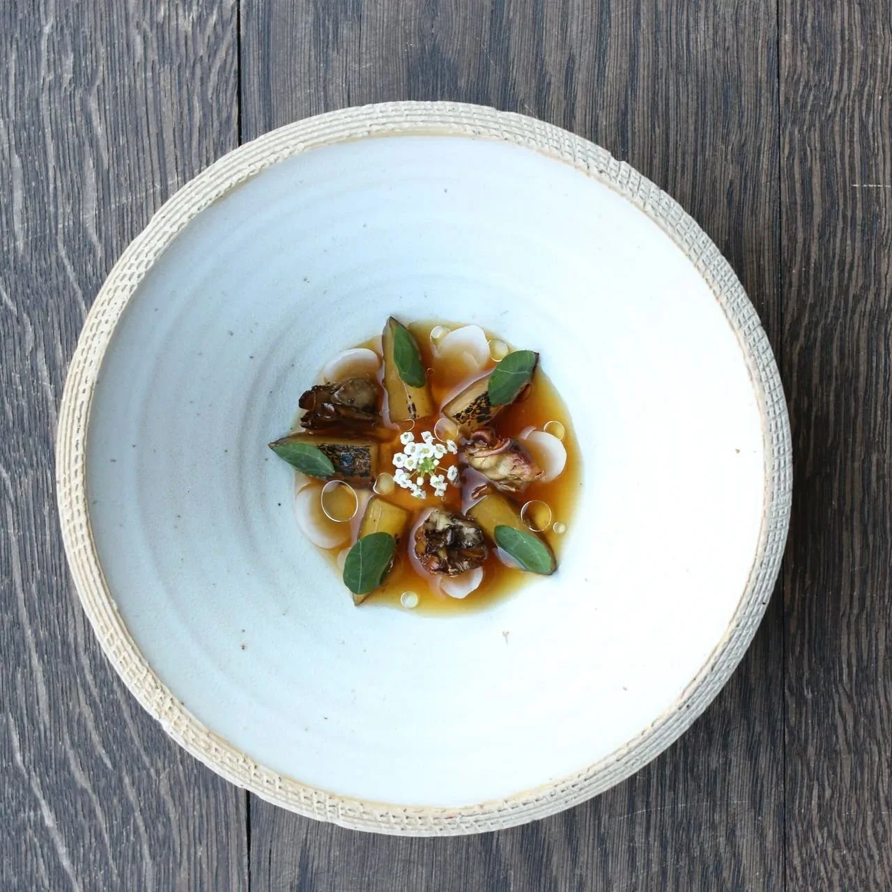 A plated gourmet dish with vegetables, herbs, and edible flowers, served in a white bowl on a wooden surface.