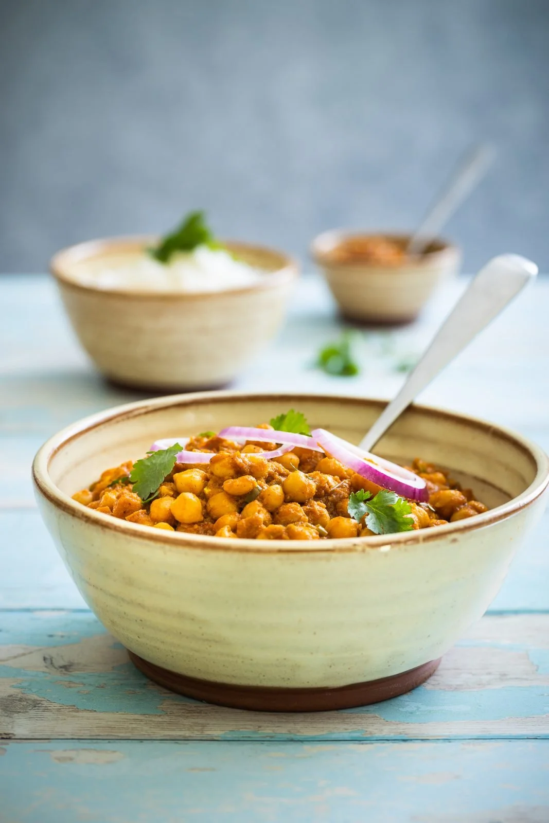A bowl of chickpea curry garnished with cilantro and red onion slices on a blue wooden table, with two additional bowls of sauces or condiments in the background.