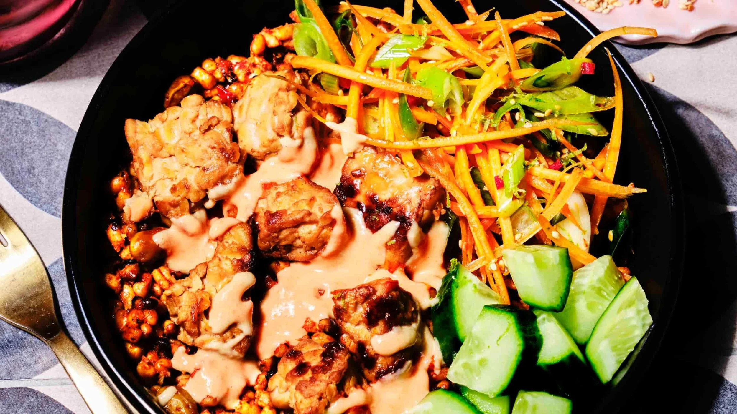 A black bowl filled with a variety of Asian-inspired dishes, including grilled chicken with a creamy sauce, shredded carrots, sliced cucumbers, and a mixed vegetable salad with sesame seeds.