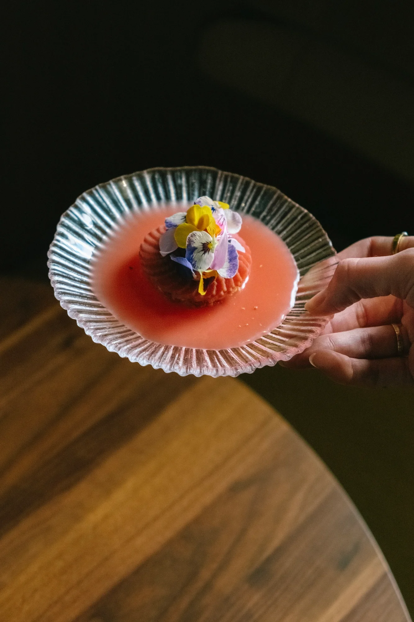 A hand holding a clear glass dish with pink-colored mousse topped with edible flowers and a cookie, placed on a wooden surface with a dark background.