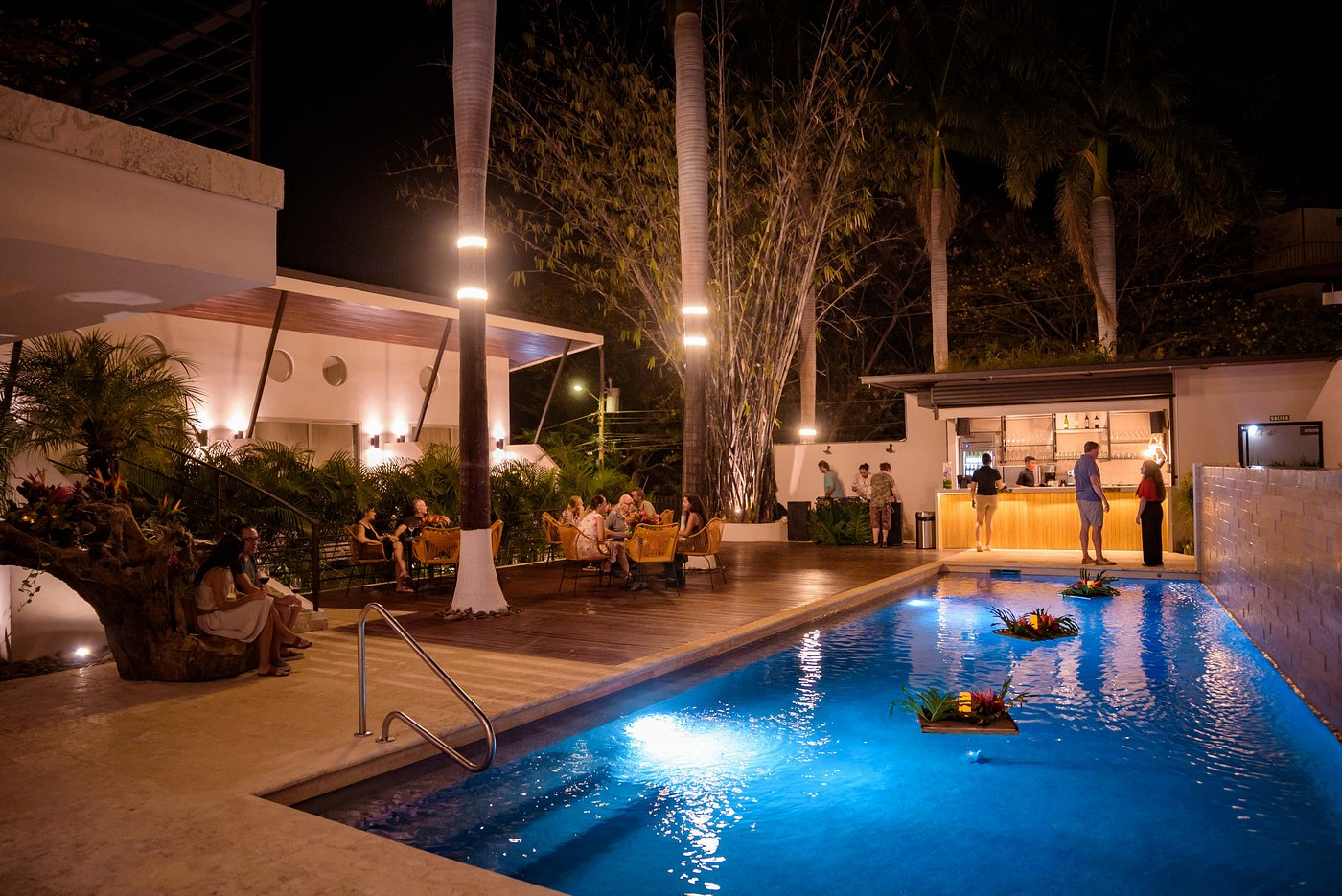 Nighttime scene of a poolside bar with people sitting at tables and ordering at the bar. The pool has floating plants with lights, and tall palm trees surround the area. There are modern buildings in the background, all illuminated with warm lighting