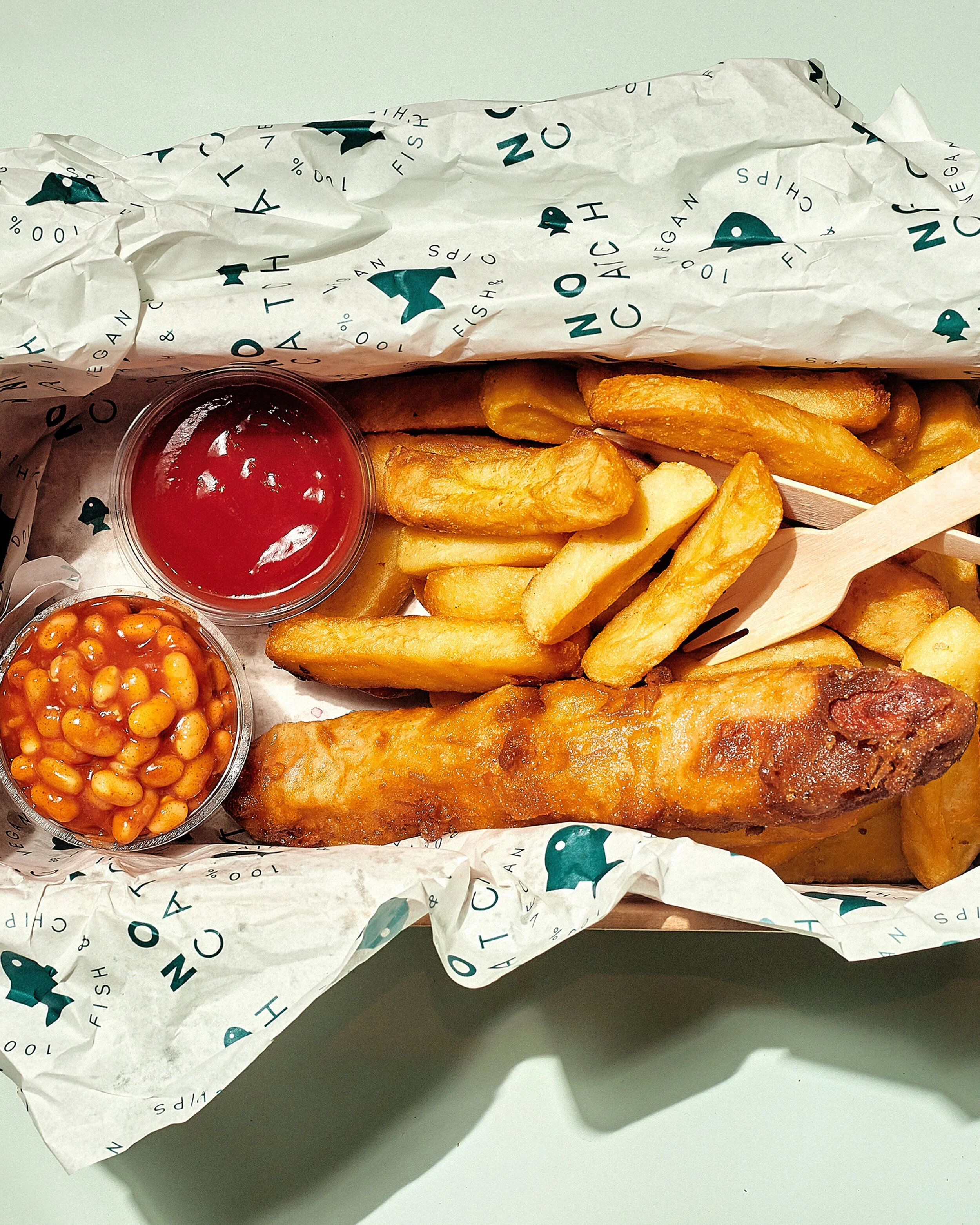 Crinkled paper wrapping with fish logo and text, partially opened to reveal fried fish fillet, thick-cut fries, a small cup of ketchup, baked beans, and a wooden fork inside a takeout container.