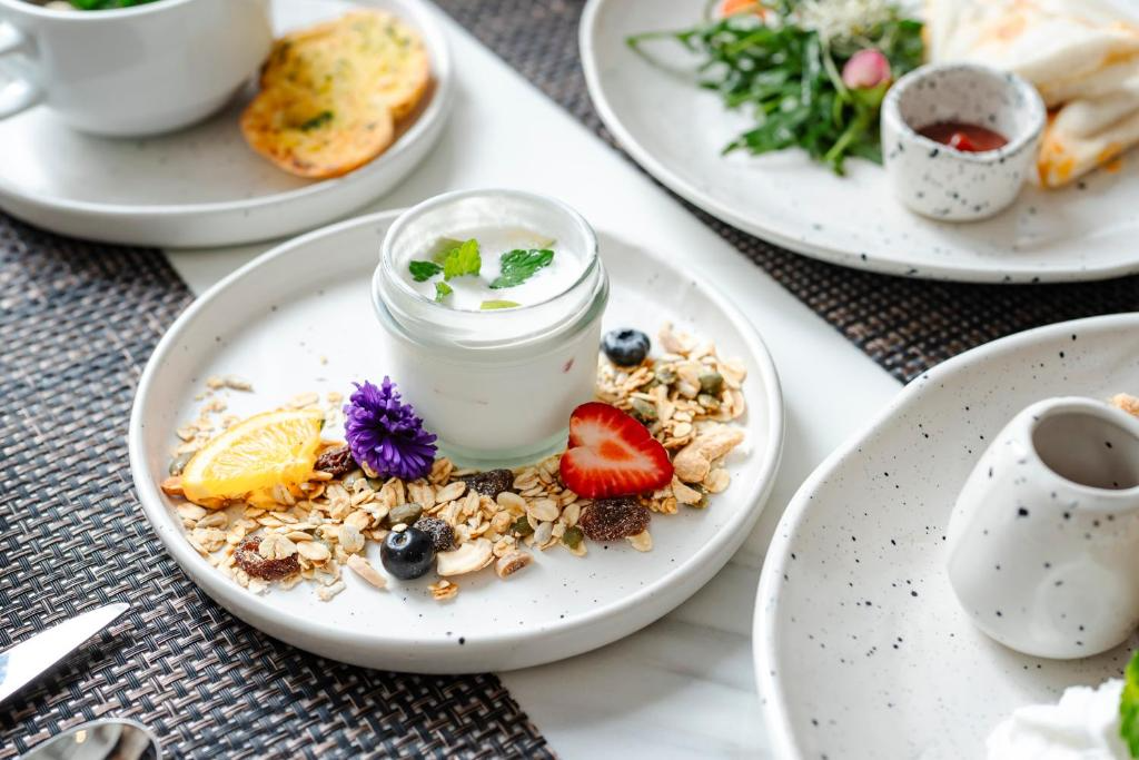 A breakfast table with a white plate holding a jar of yogurt topped with a mint leaf, a slice of lemon, a strawberry, blueberries, and granola with a purple flower decoration.