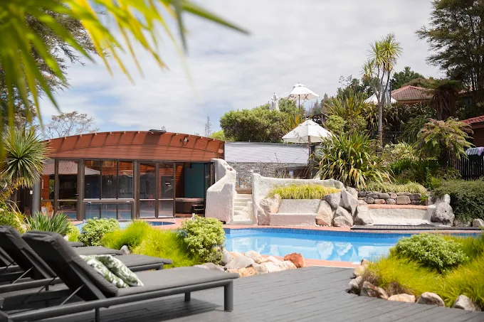 A backyard swimming pool with lounge chairs, greenery, rocks, and a covered patio area with umbrellas.