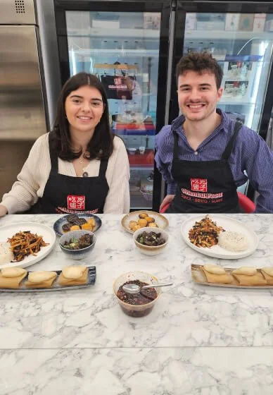 Two smiling people wearing aprons standing behind a marble counter with various prepared dishes in front of them, including wraps, bowls of food, and a small pot of sauce, in a kitchen or restaurant setting.