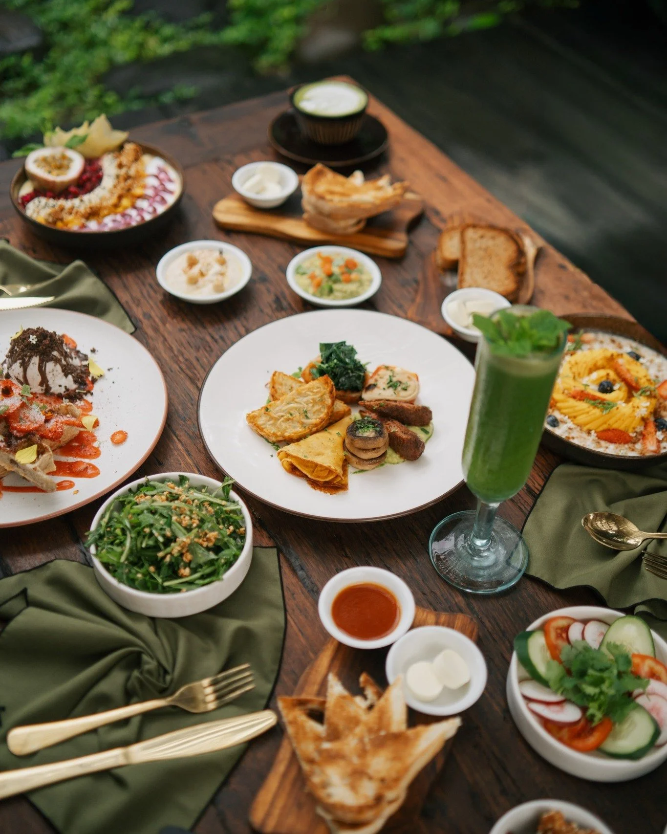 A variety of dishes on a wooden table, including salads, a green drink, bread, and desserts, with green napkins and gold utensils.