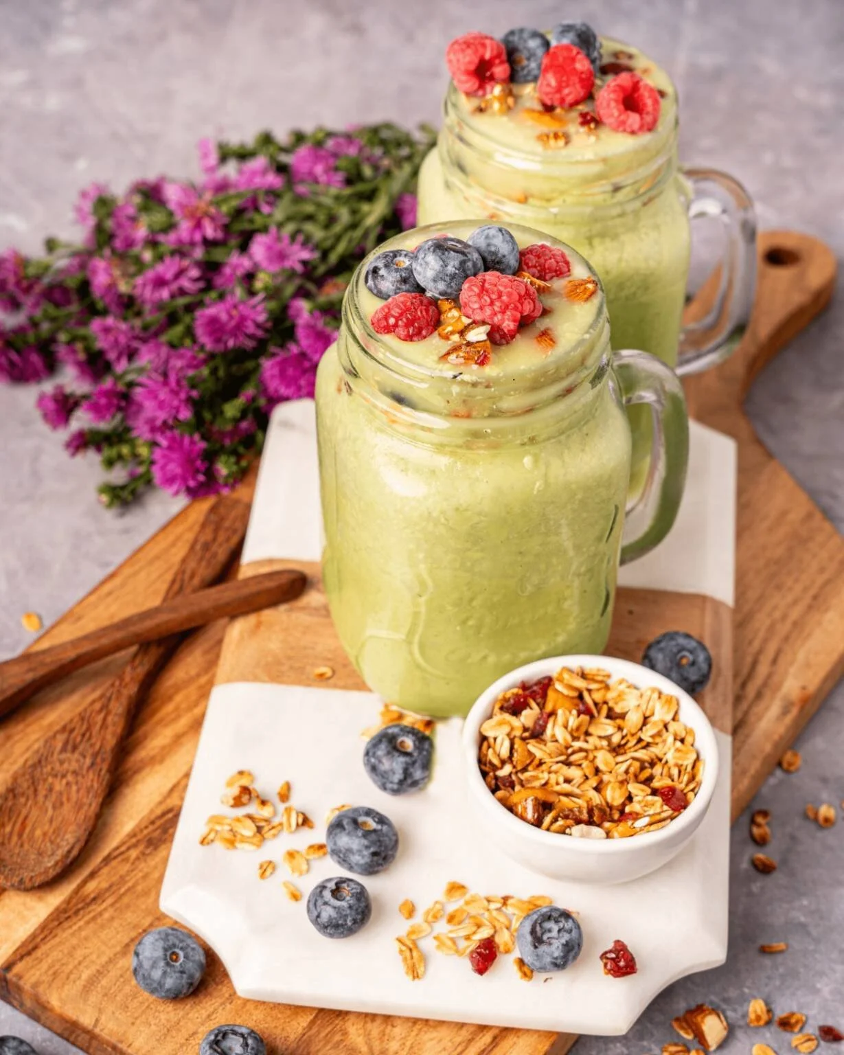 Two mason jar smoothies topped with mixed berries and granola, placed on a wooden serving board with a small bowl of granola, blueberries scattered around, and a bunch of purple flowers in the background.