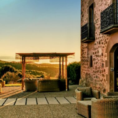 Outdoor patio with wicker furniture under a wooden pergola, overlooking a scenic sunset view with trees and open landscape.