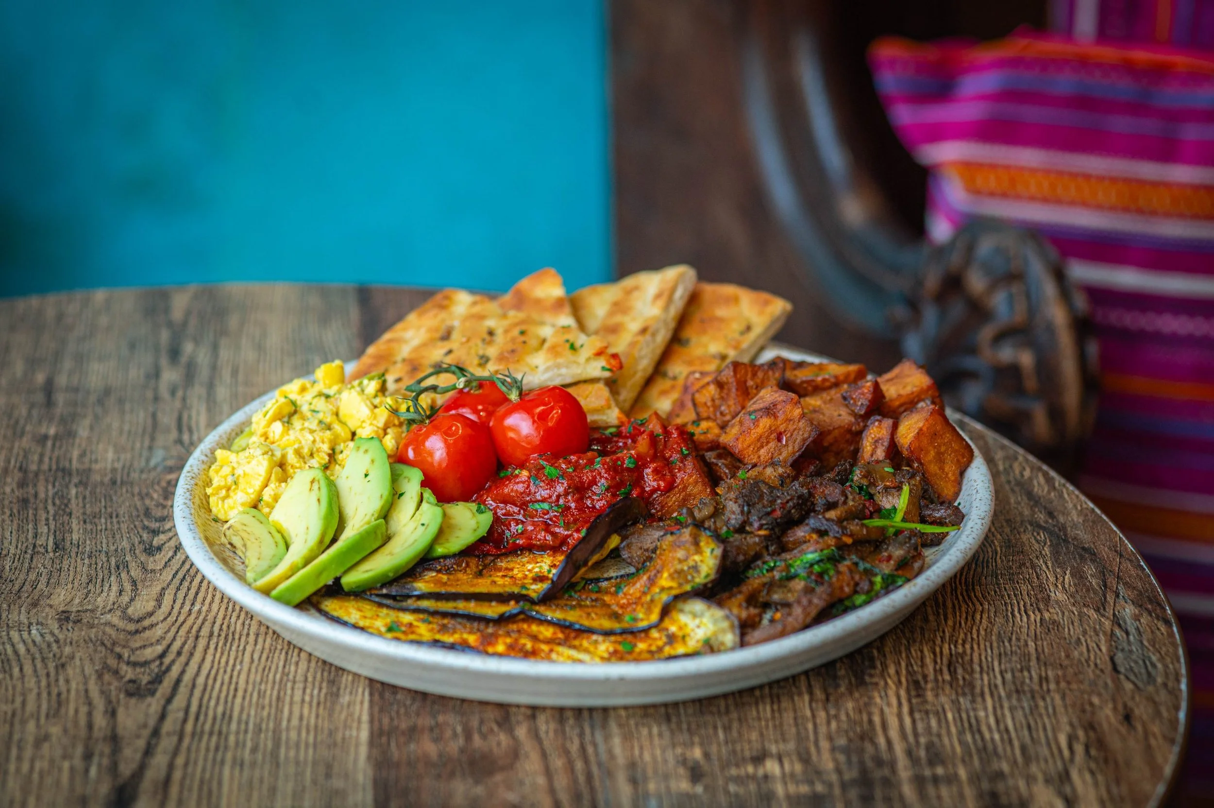A large, oval ceramic plate filled with various Mexican dishes, including sliced avocado, scrambled eggs with herbs, red tomatoes, layered tortillas with cheese, grilled meats, and stewed vegetables, set on a wooden table.
