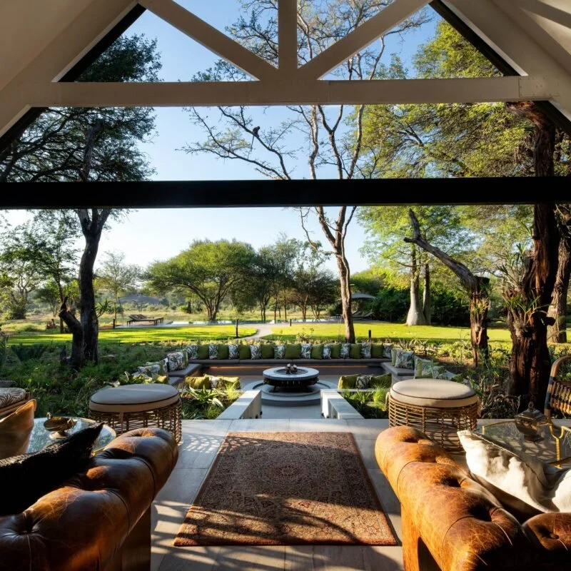 Interior view of a spacious living room opening to a lush outdoor garden with trees and a grassy lawn, featuring stylish leather and wicker furniture, decorative cushions, and a central fire pit.