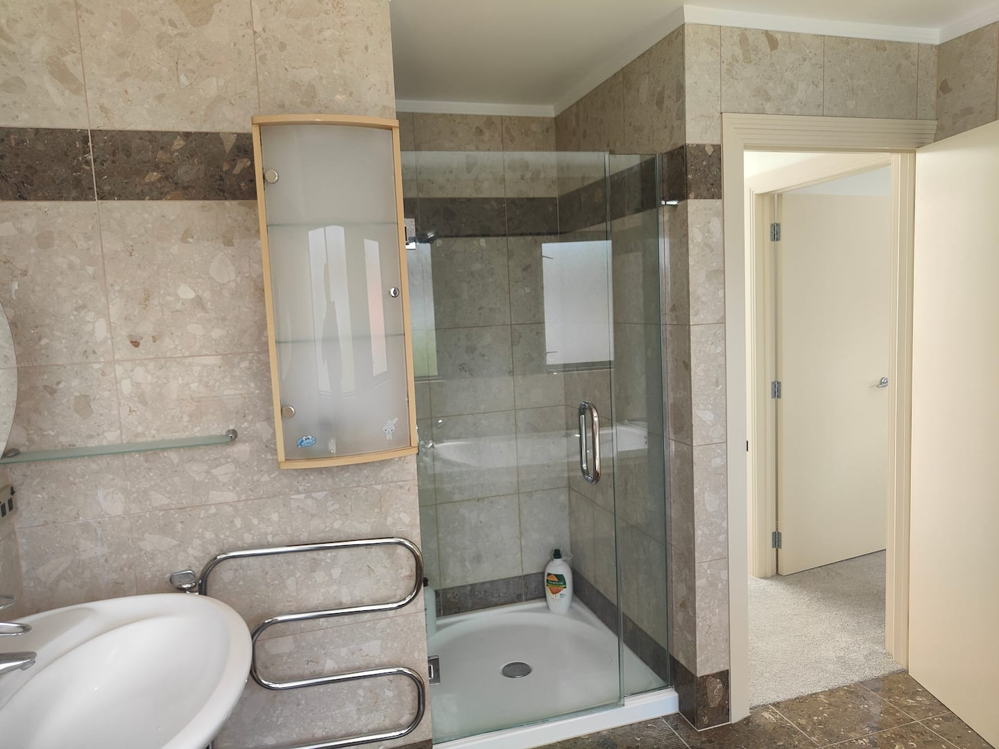 A bathroom with beige tiled walls and a glass shower enclosure. There is a small frosted glass cabinet above the shower and a white sink with a chrome towel rack below. An open door leads to a room with carpeted flooring.
