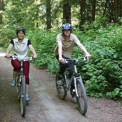 Two women riding bikes on a wooded trail surrounded by lush greenery.