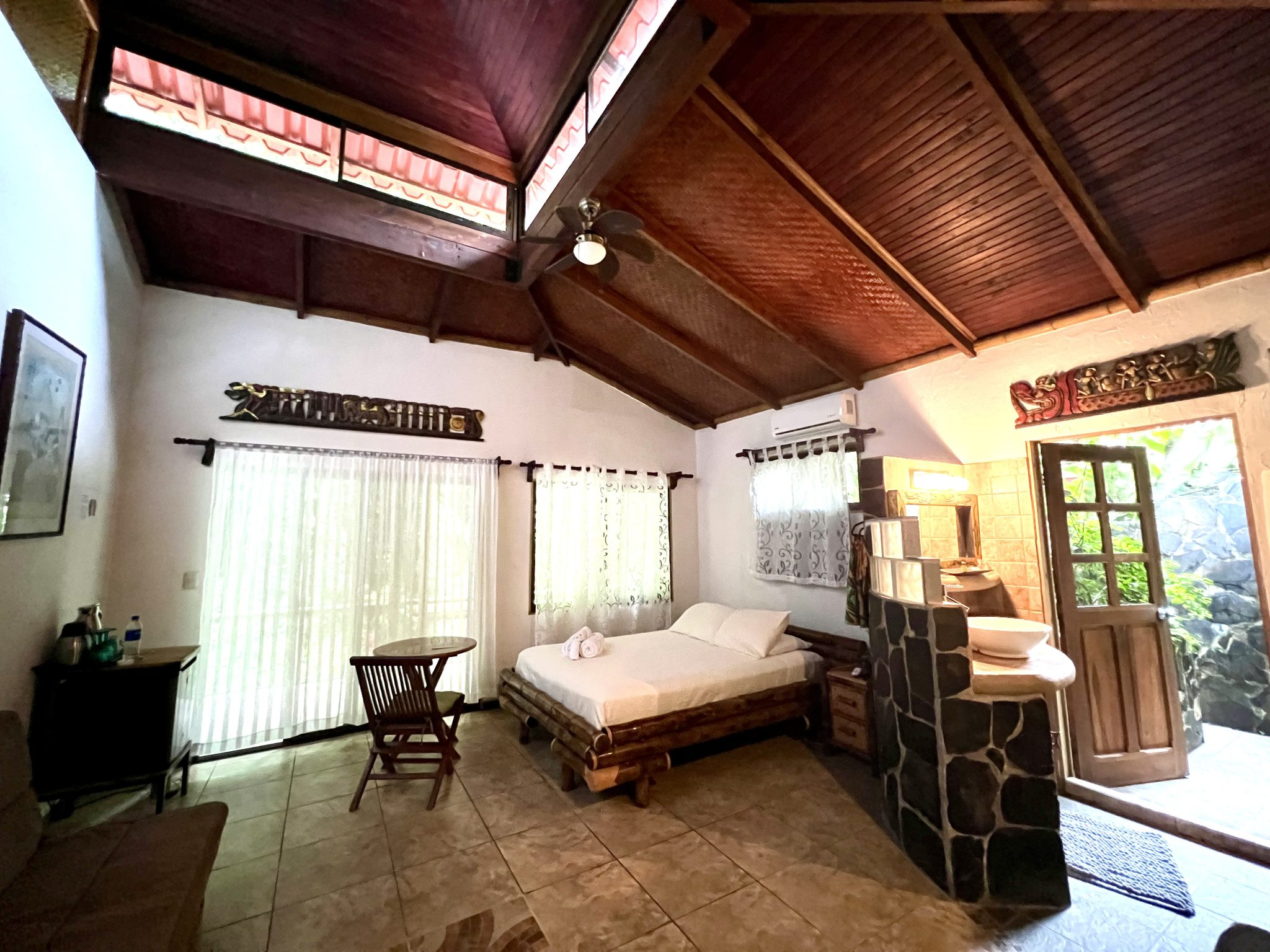 Living room with a wooden ceiling, white walls, and large windows with curtains. There is a wooden bed with a mattress, a small table, and an air conditioning unit on the wall. The room has tropical decorative elements and tiled floors.