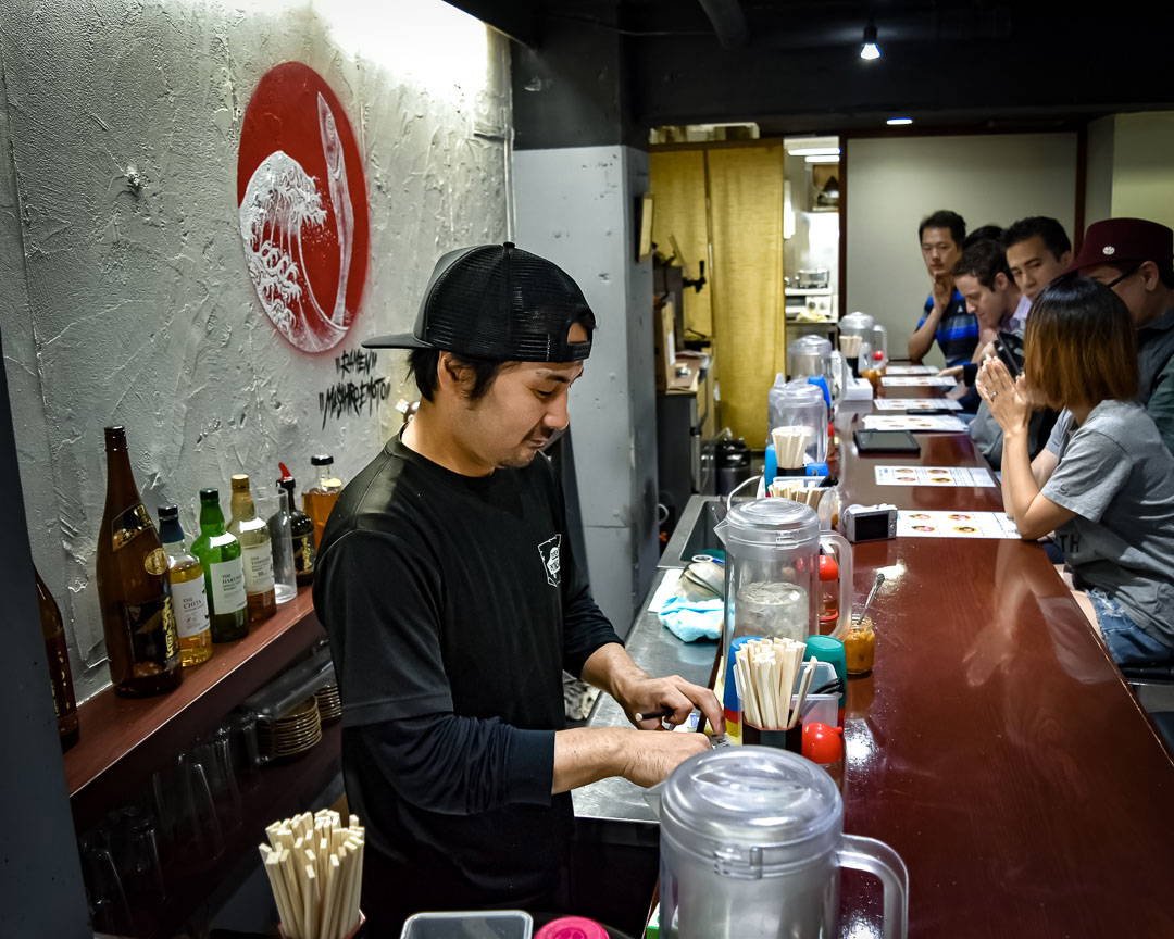 A chef in a black shirt and cap preparing food at a counter in a Japanese restaurant, with customers seated along the bar.